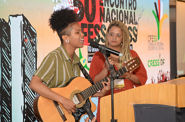 Imagem mostra as duas assistentes sociais em pé, fazendo apresentação artística no evento, à frente do telão de led com a arte do Encontro Nacional..