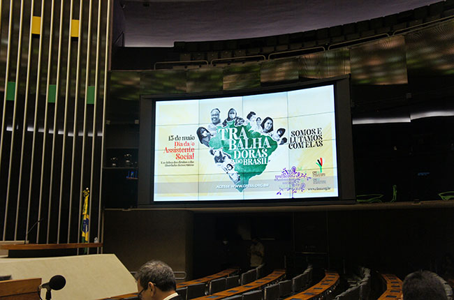 Imagem de dentro do Plenário da Câmara dos Deputados mostra um telão, no qual está a imagem do cartaz do Dia da Assistente Social 2022.