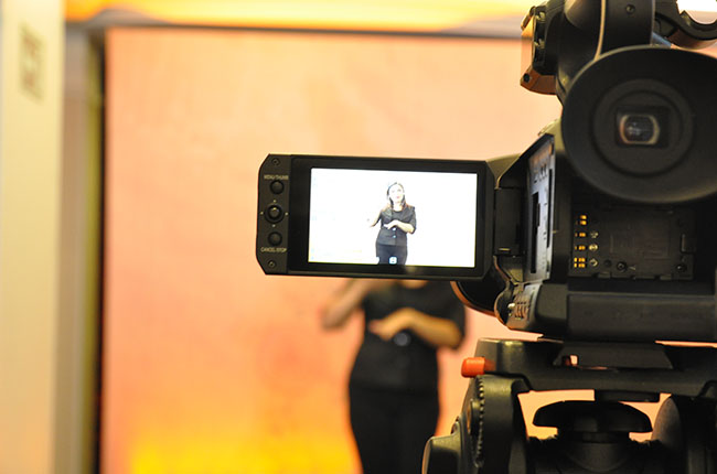Imagem mostra uma câmera de filmagem e, na tela, a profissional intérprete de Libras no estúdio. 