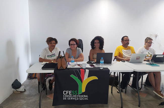 Imagem mostra as representantes do CFESS no FNRU sentadas à mesa da coordenação, juntamente com a presidenta do CRESS-MT.