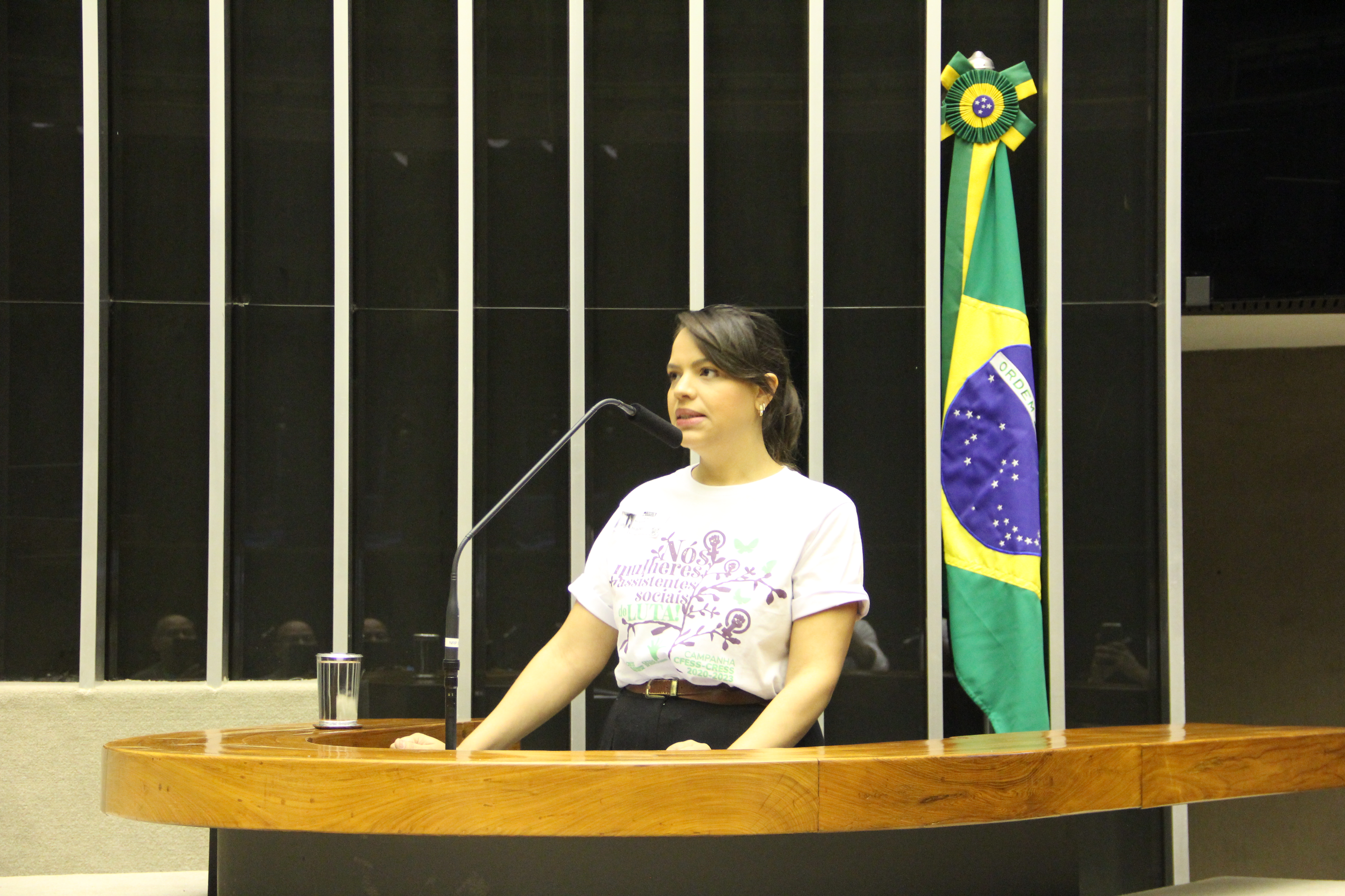 Imagem mostra a conselheira do CFESS Rafaella da Câmara, em pé na tribuna, com uma camiseta branca e a bandeira do Brasil ao fundo. 