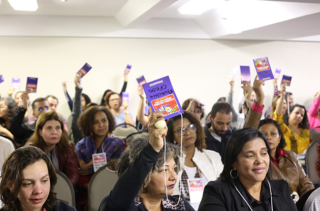Imagem mostra plenária de assistentes sociais, com braços para cima estendendo seus crachás, para aprovar o voto on line para a categoria
