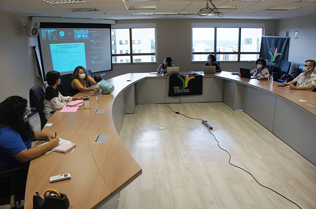 Imagem mostra uma mesa central, com representantes da gestão do CFESS ao redor. No canto superior esquerdo, um telão mostra outros membros da diretoria em participação online. 
