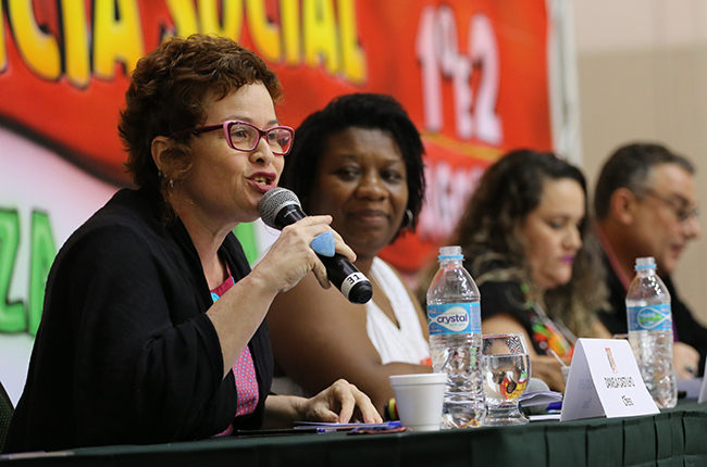 Foto mostra a palestrante Daniela Castilho de perfil, ao lado de outras que compuseram a mesa, palestrando sobre a Agenda Política da categoria