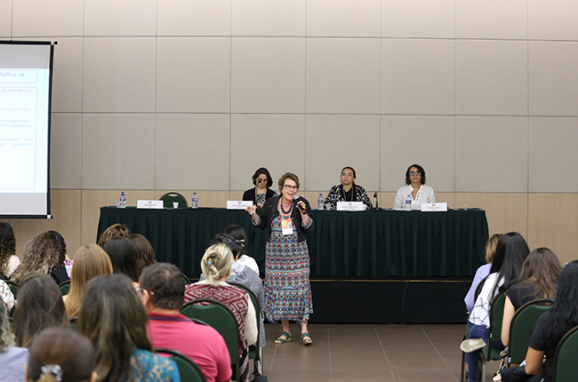Foto mostra a palestrante Regina Mioto em pé apresentando slides para o auditório cheio de pessoas
