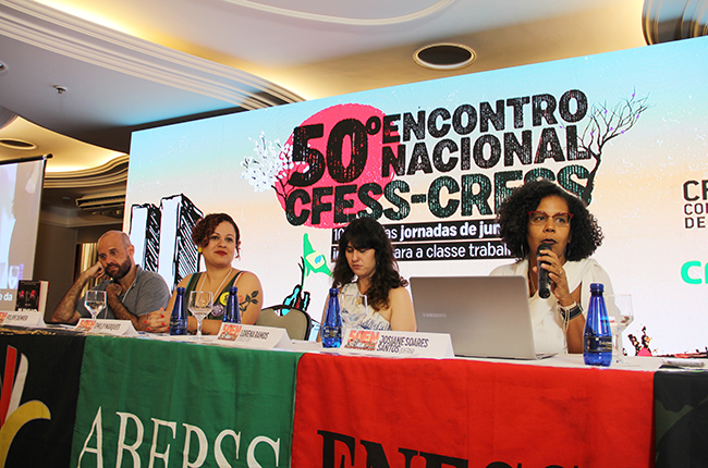 Imagem mostra as 4 pessoas sentadas compondo a mesa da conferência de abertura do evento, com o telão de led ao fundo e a professora Josiane Soares falando.