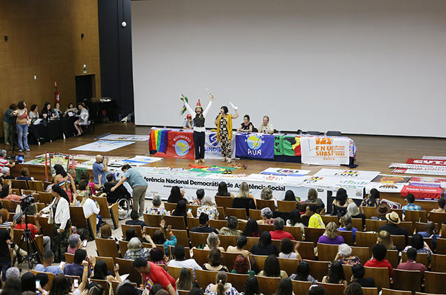 Imagem mostra o auditório lotado durante a conferência, com a mesa coordenadora em cima do palco e a conselheira do CFESS Elaine Pelaez em sua fala.