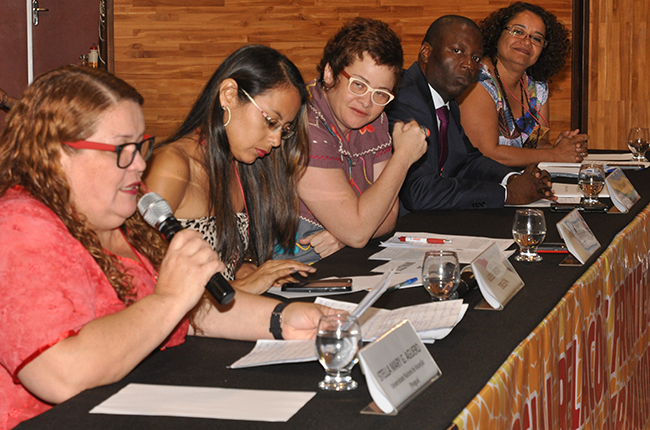 Foto com imagem em destaque de Stella Aguero (do Paraguai). Na outra ponta da mesa Esther Lemos (CFESS) e Simão Samba (Angola) 