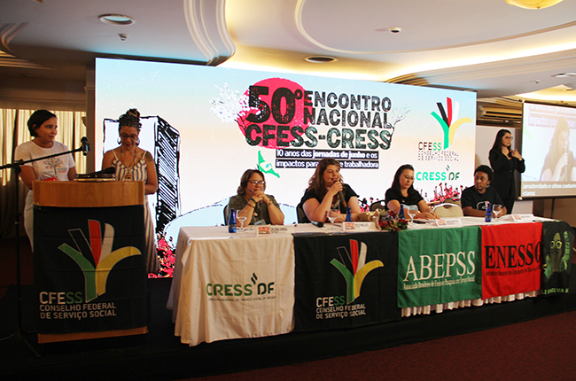 Imagem mostra a mesa de abertura do evento, com palestrantes sentadas, a presidenta do CFESS falando ao microfone e duas mulheres em pé no púlpito à esquerda.