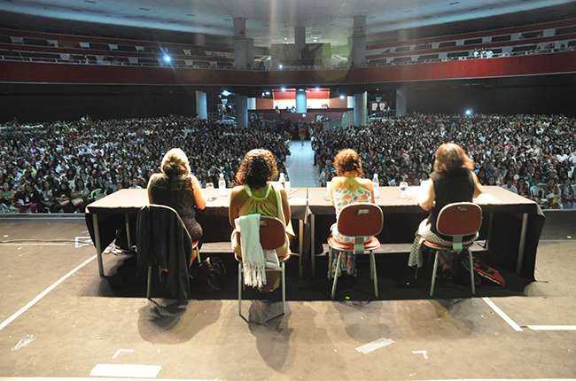 Imagem mostra vista panorâmica do palco onde estava as palestrantes da conferência de abertura e o público ocupando grande parte do local  