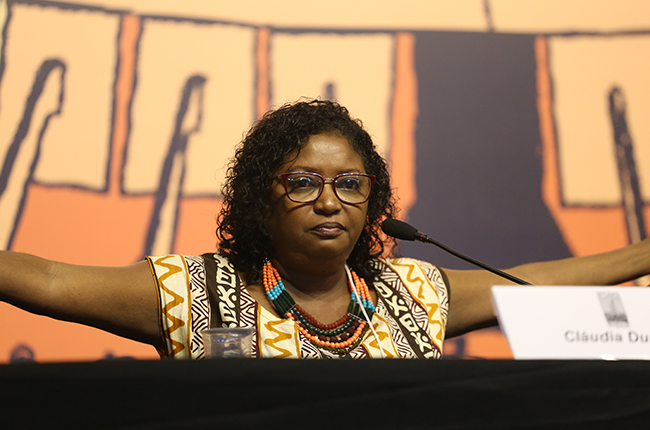 Foto de Cláudia Durans, mulher negra que está de braços em frente ao público para reafirmar a resistência do povo negro