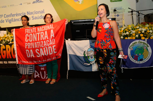 Imagem mostra a conselheira do CFESS Elaine Pelaez durante tribuna na Conferência. Ao fundo, duas mulheres seguram a faixa da Frente Nacional Contra a Privatização da Saúde