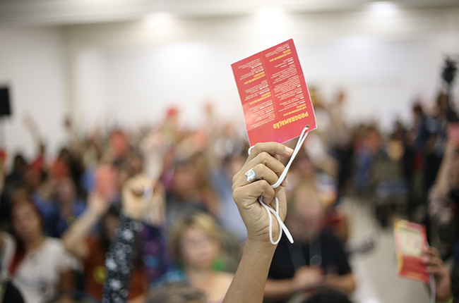 Imagem mostra mão de mulher em destaque, segurando o crachá vermelho de delegada no momento da votação. Ao fundo da imagem está o restante da plenária, desfocado.