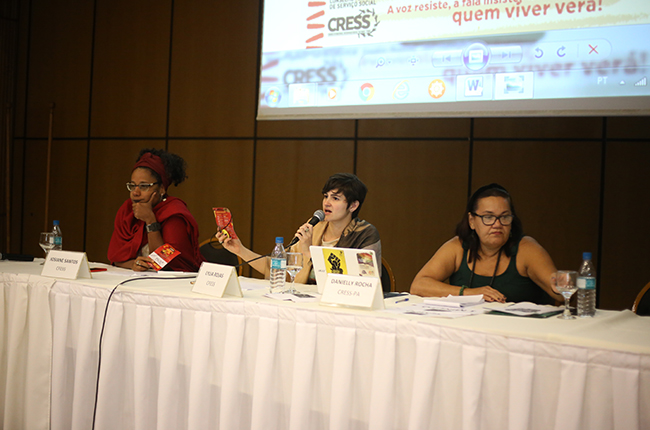 Foto mostra as integrantes da mesa que conduziram a Plenária Final. Nela, as integrantes estão segurando o microfone para dar orientações aos/às participantes