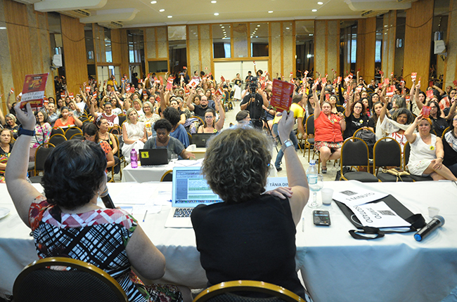Imagem mostra foto da plenária, momento em que os/as participantes levantam seus crachás vermelhos aprovando as mudanças no Código Eleitoral