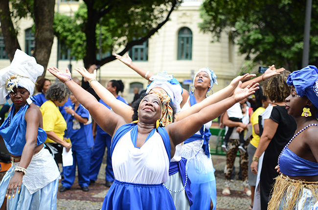 Imagem mostra dança do grupo Filhas de Ghandy. As mulheres negras estão de roupa azul e branco, olhando para o céu, de braços abertos.