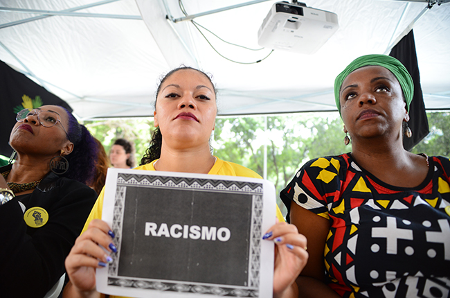Imagem mostra mulheres negras assistindo à aula pública sobre racismo, na tenda montada pelo CFESS e CRESS-RJ. Uma delas segura o cartaz escrito a palavra racismo