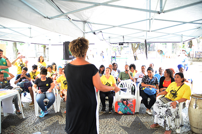 Imagem mostra várias pessoas sentadas, sob a tenda montada para a atividade, assistindo à aula da assistente social Roseli Rocha sobre racismo. 
