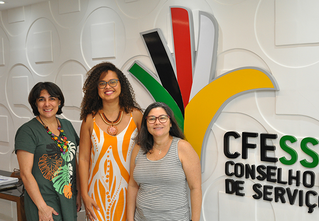 Imagem mostra as 3 integrantes da CNE em pé, em frente ao letreiro com o nome do CFESS na sede da entidade.