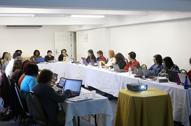 Foto mostra as conselheiras do CFESS e as assessorias sentadas em uma mesa em forma da letra U, debatendo sobre os eventos do CFESS