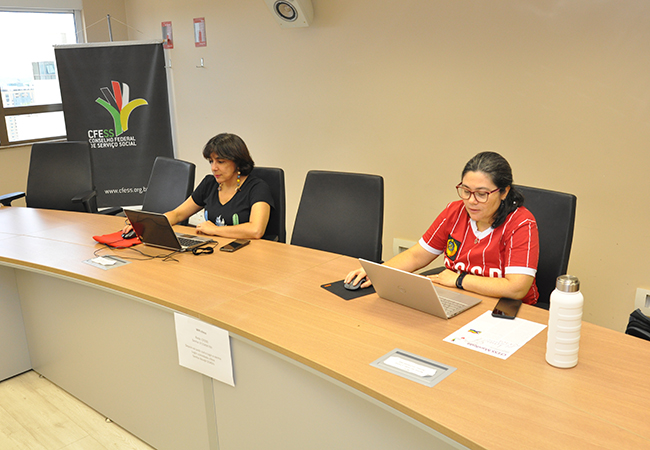 Imagem mostra duas mulheres sentadas, trabalhando no computador na sede do CFESS, com o banner do CFESS ao fundo. 