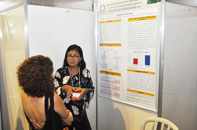 Imagem mostra a assistente social Ana Cláudia Martins falando sobre sua pesquisa para uma participante do evento.