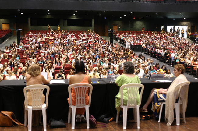 Imagem do alto do palco do auditório do centro de convenções mostra a mesa com palestrantes e o público ao fundo.