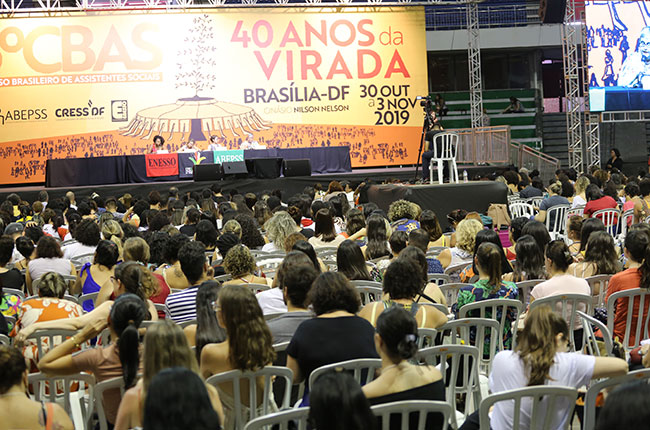 Imagem do público assistindo à mesa da tarde, com o palco e as palestrantes ao fundo.