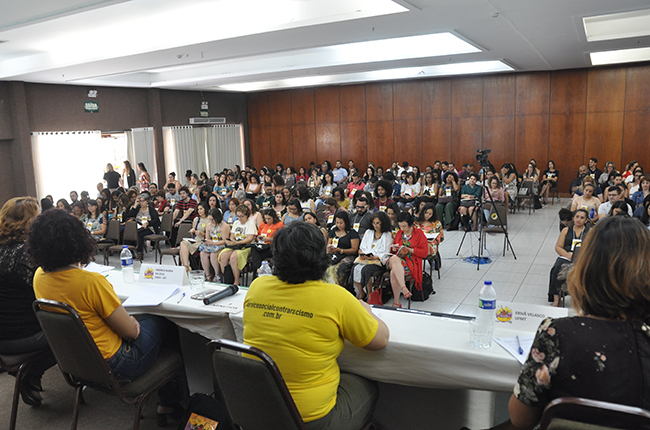 Fotografia tirada na perspectiva atrás das palestrantes mostra auditório cheio, com participantes acomodados/as nas cadeiras