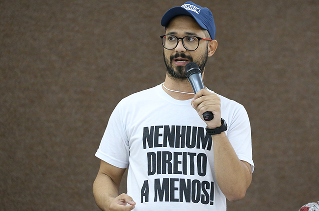 Imagem mostra o assistente social Fábio Santos (Sintufs) em pé, segurando microfone em sua fala. Ele está com uma camisa com os dizeres nenhum direito a menos