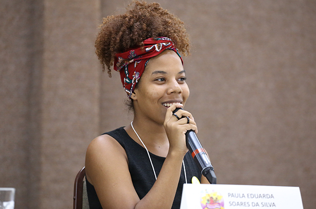 Fotografia mostra Paula Soares, estudante de Serviço Social da UFMT, falando para o público. Sua expressão é de alegria