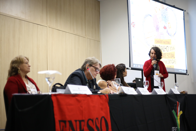 Imagem mostra a palestrante Rosa Prédes (Ufal) em destaque na mesa de debates, enquanto outros/as palestrantes acompanham sua fala