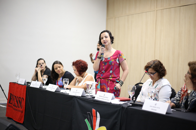 Imagem mostra participantes da mesa de abertura. Em destaque a conselheira do CFESS Elaine Pelaez com microfone na mão