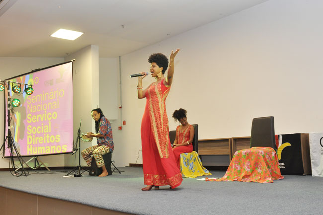 Imagem da atividade cultural ao fim do primeiro dia, com apresentação do Recital Vozes Negras, com três mulheres negras e dois músicos no palco. 