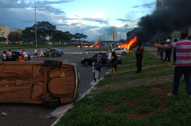 Foto mostra carro em chamas e outro virado. Protesto de manifestantes frente à violência da PM