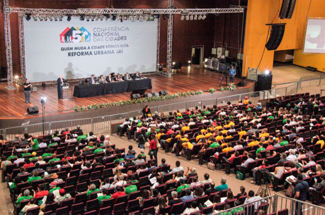 Imagem mostra auditório visto de cima e participantes da Conferência