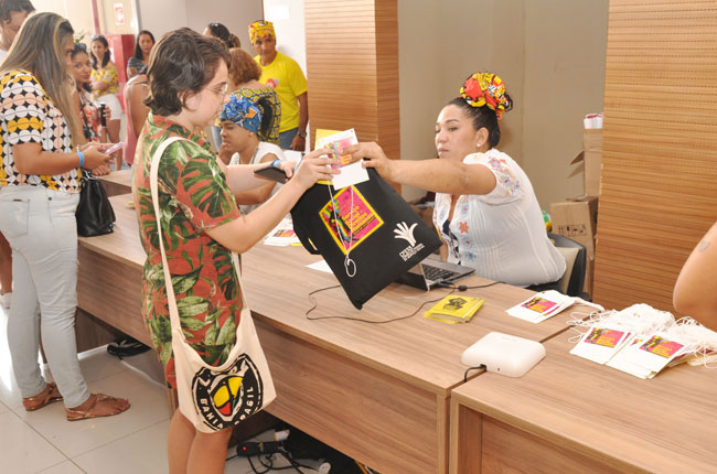 Imagem do credenciamento do evento, no momento em que uma recepcionista entrega o material a uma participante. 
