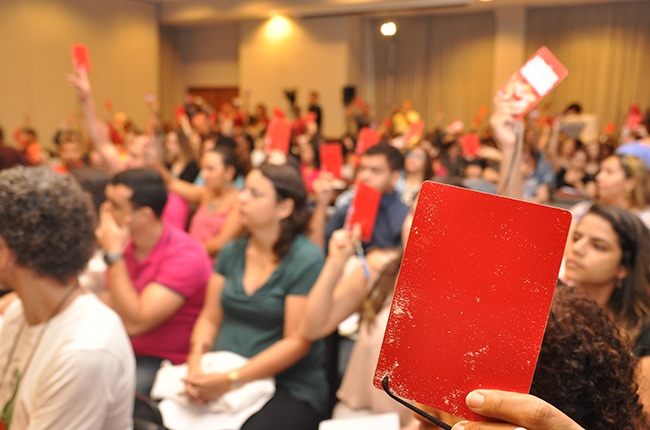 Imagem mostra assistente social levantando um crachá vermelho, de delegado(a), durante a votação na Plenária Final